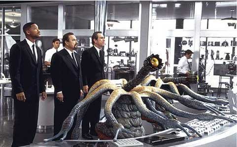 Agents J, K and Zed stand in the centre of the MiB head quarters surrounded by various lab coats with experimental weapons on the walls; in front of them are the alien twins working away at a console: they are tallish creatures with an octopus leg like top which through the centre is a long stalk with a yellow eyeball on the end